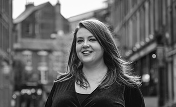 Black and white photo of young White woman with long brown hair, smiling, in city street setting 