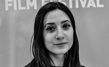 Black and white image of young white woman with long dark hair standing in fro of film festival poster