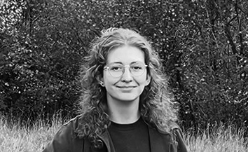 Black and white head and shoulders shot photo of young White woman with long curly hair and wearing glasses in rural outdoor setting.