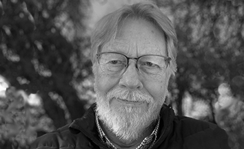 Black and white head and shoulder shot of white man with grey hair and beard and glasses in outdoor location