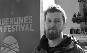 Black and white head and shoulder shot of young white man with bears standing close to a Borderlines Film Festival banner