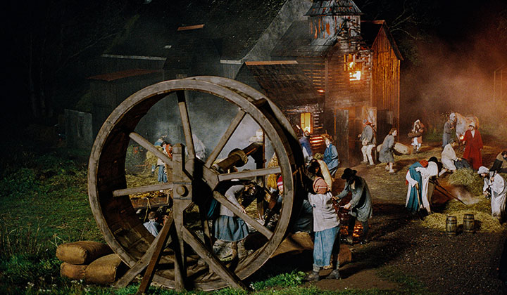 Night shot with wooden mill wheel in foreground and burning building in background. Many characters scuttling around between the two.