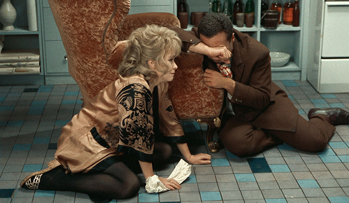 woman with blonde hair swept upon a chignon kneeling on the floor , man win suit with his face buried in the arm of a chair as though in distress