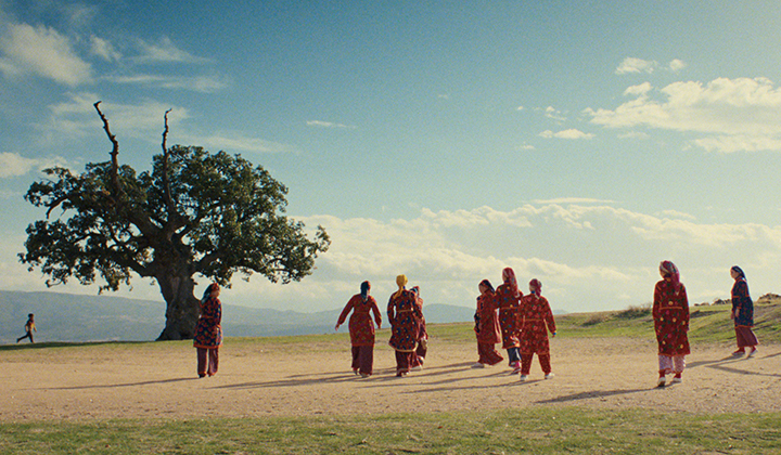 Women dressed in red traditional costume playing football in sunny, dry landscape, tree on left and mountains in the distance