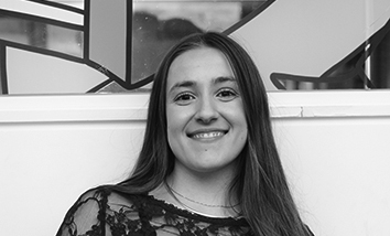 Black and white image of a young white woman with long, straight dark hair in front of a patterned background.