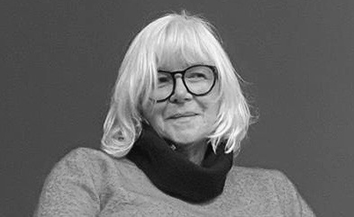 White woman with shoulder-length white hair with fringe, wearing dark-rimmed glasses. Dark background.