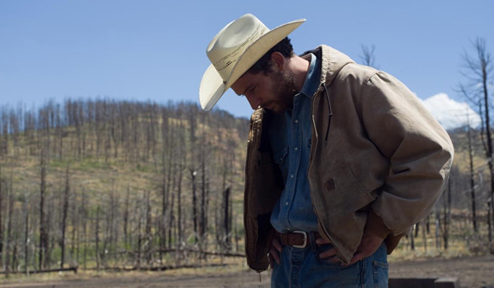 Man in cowboy hat looking down, background landscape of burned trees