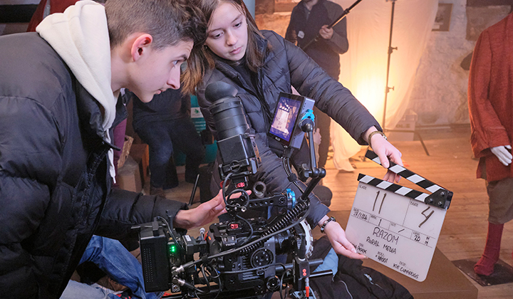 Young man in profile holding camera, young woman in background holding clapperboard, light behind her.