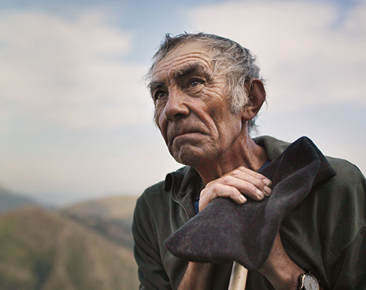 Older man with hands leaning on a stick or crook looking to one side, mountain landscape.