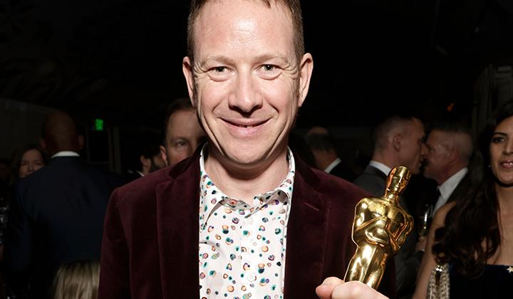 Middle-aged white man smiling, holding Oscar award. Wearing patterned shirt and dark jacket, dark background with people visible.