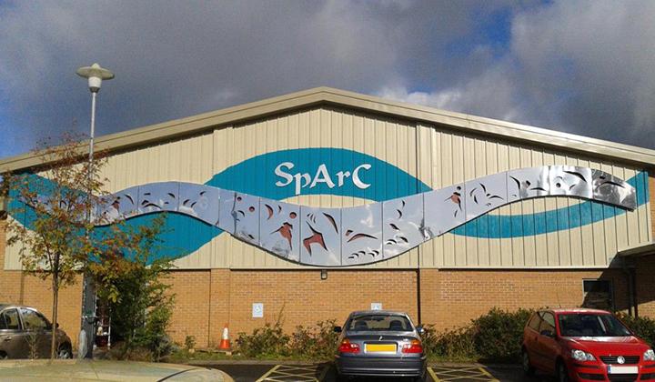 Large arts/leisure centre with a sloping roof. Lower part of building is brick, upper part metal clad with a wavy turquoise and metal banner.