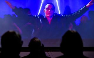 Man with outstretched arms on blue background, heads of audience watching in foreground 