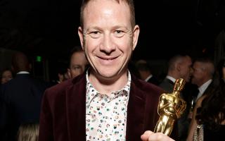 Middle-aged white man smiling, holding Oscar award. Wearing patterned shirt and dark jacket, dark background with people visible.