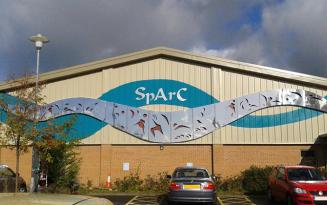 Large arts/leisure centre with a sloping roof. Lower part of building is brick, upper part metal clad with a wavy turquoise and metal banner.