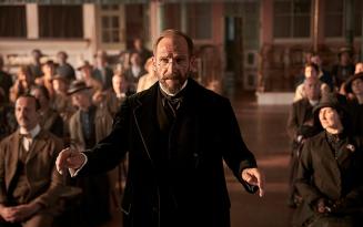 Older man in dark clothes standing in front of a hall falloff seated people