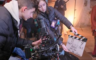 Young man in profile holding camera, young woman in background holding clapperboard, light behind her.