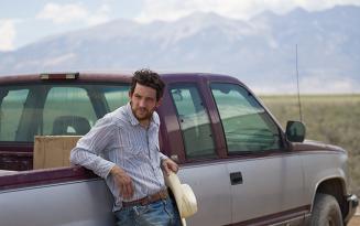 Young white man with beard in blue and white striped shirt leaning against a pick up truck, mountains in the distant background