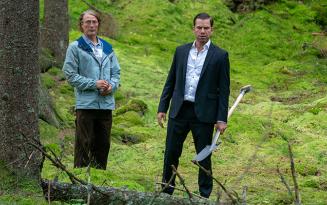 Two mention woodland setting, one in a suit carrying a long spade, the other with a blond perm and glasses in casual clothes ,