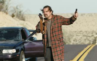 man wearing checked brown dressing gown holding a phone with one arm raised by a car on an open road