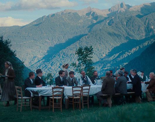 People, mainly men, seated outside at a table with a back drop of high green mountains