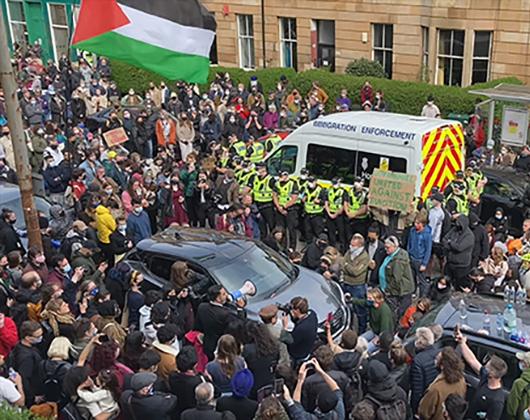Crowd of people in city street surrounding a police van, Palestinian flag in one corner