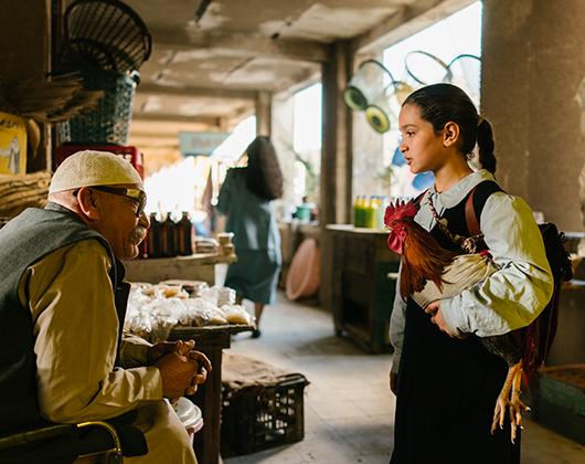 girl in school pinafore with white blouse holding rooster in an indoor market